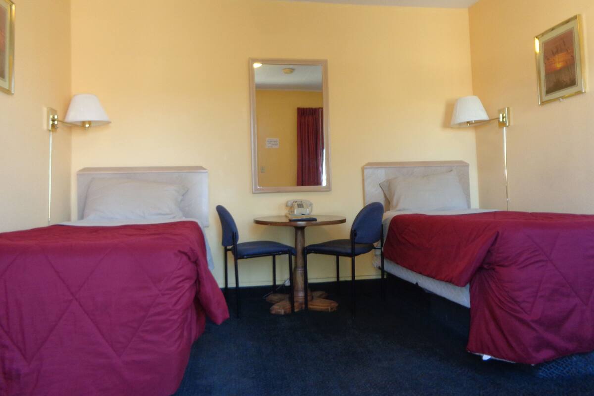 Camera d'albergo con due letti singoli, un tavolino, due sedie, uno specchio e due lampade.