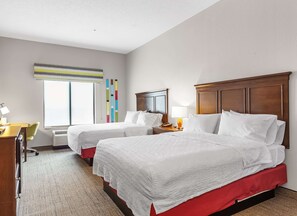 A hotel room with two beds, a desk, a chair, a lamp, and a window with blinds.