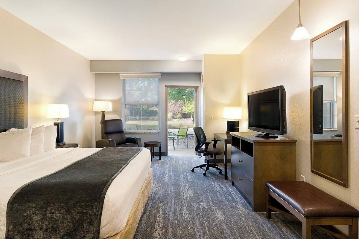Ein Hotelzimmer mit einem großen Bett, einem Schreibtisch mit Fernseher, einem Sessel, einer Bank und einem großen Fenster mit Blick ins Freie.