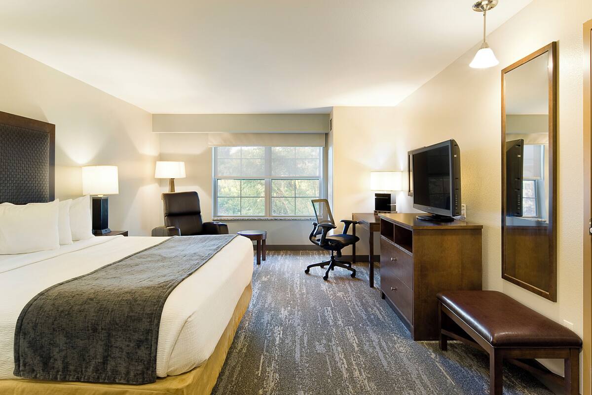 Ein Hotelzimmer mit einem großen Bett, einem Schreibtisch, einem Sessel, einem Fernseher, einer Bank und einem Fenster.
