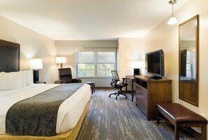 Quarto de hotel com uma cama grande, uma escrivaninha com computador, televisão e janela com vista para árvores.