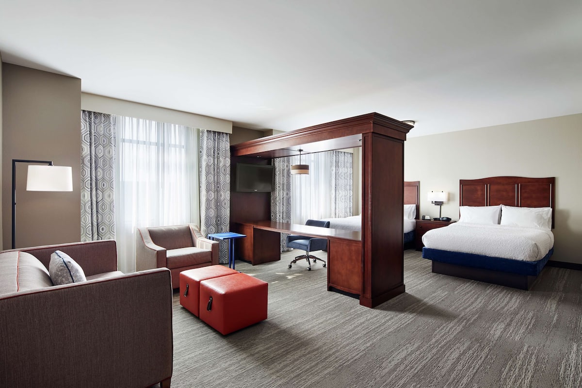 A hotel room with a bed, a desk, a chair, a sofa, and a small red ottoman.