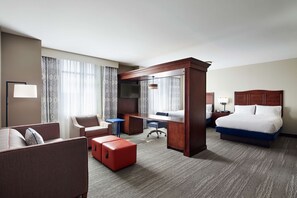A hotel room with a bed, a desk, a chair, a sofa, and a small red ottoman.