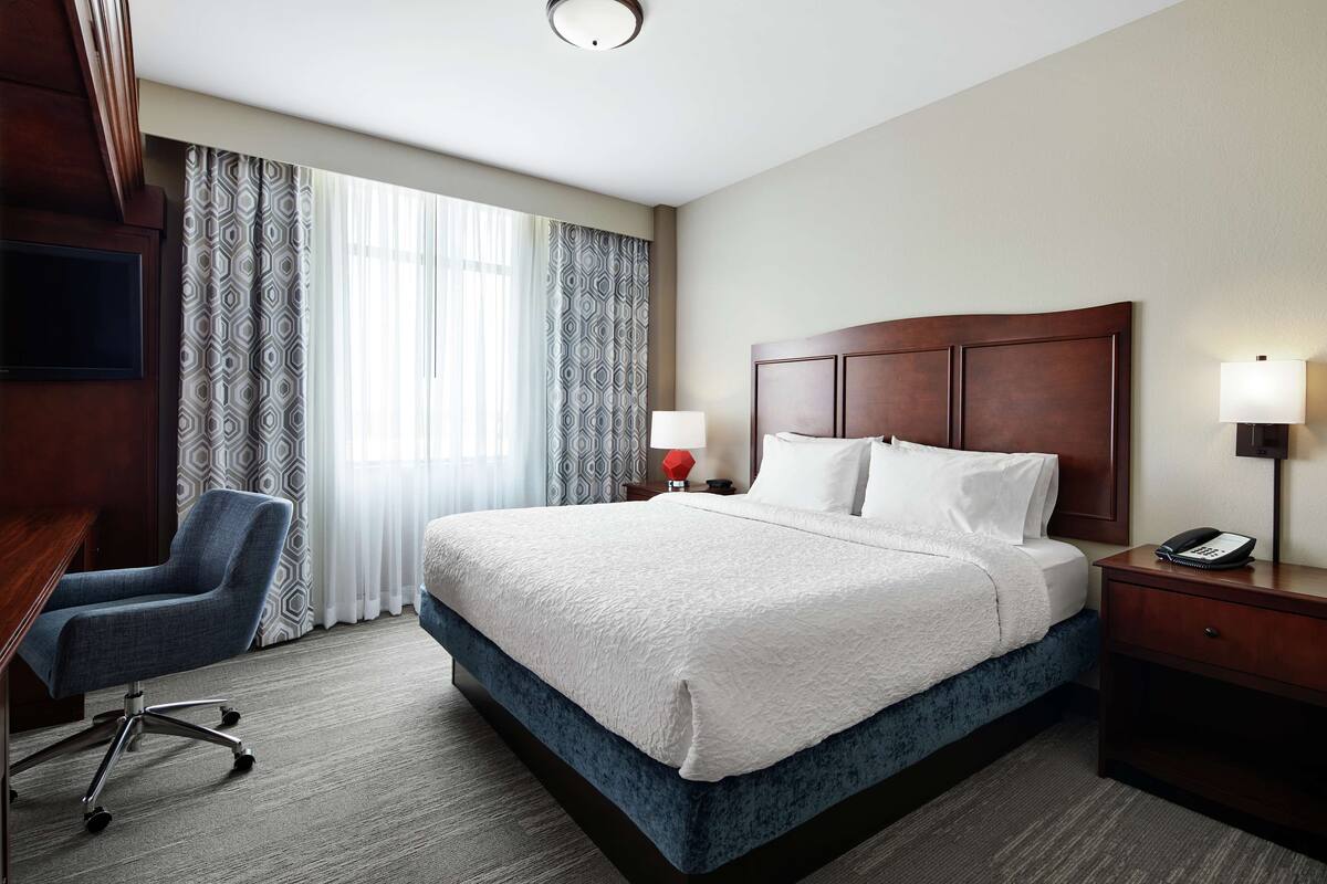 A hotel room with a large bed, a desk, a chair, a nightstand with a lamp, and a TV.
