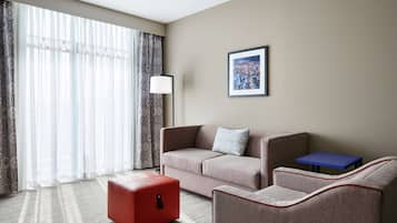 A hotel room with a sofa, a red ottoman, a small table, and a lamp.