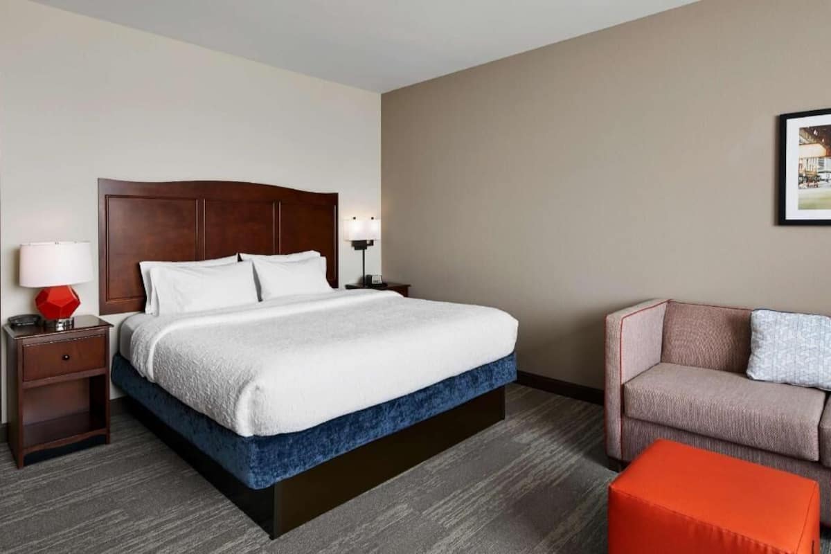 A hotel room with a large bed, a nightstand with a lamp, a sofa, and an ottoman.