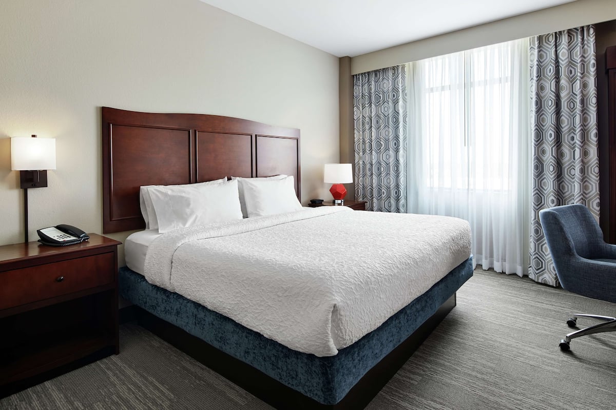 A hotel room with a large bed, two bedside lamps, a nightstand with a phone, a chair, and a window with patterned curtains.