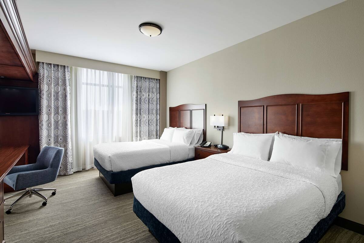 A hotel room with two beds, a desk, a chair, a TV, and a window with curtains.