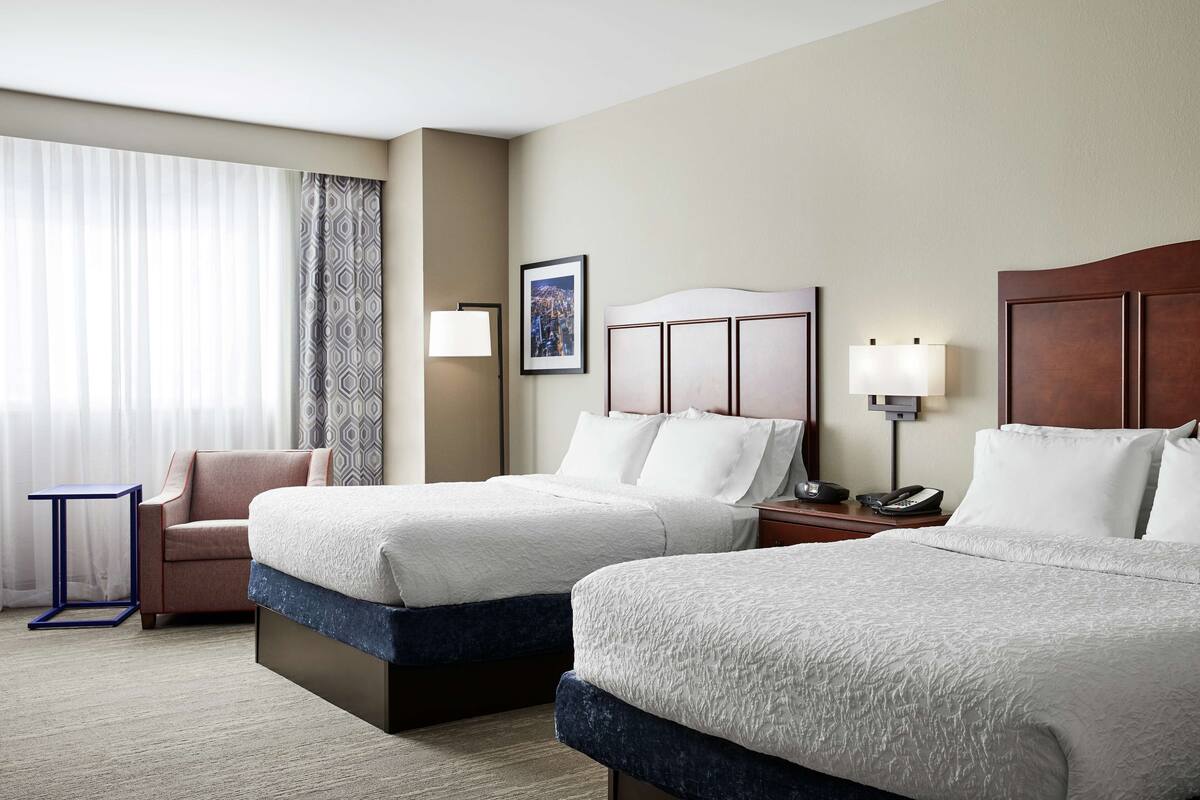 A hotel room with two beds, a chair, a small table, and a nightstand with a lamp.