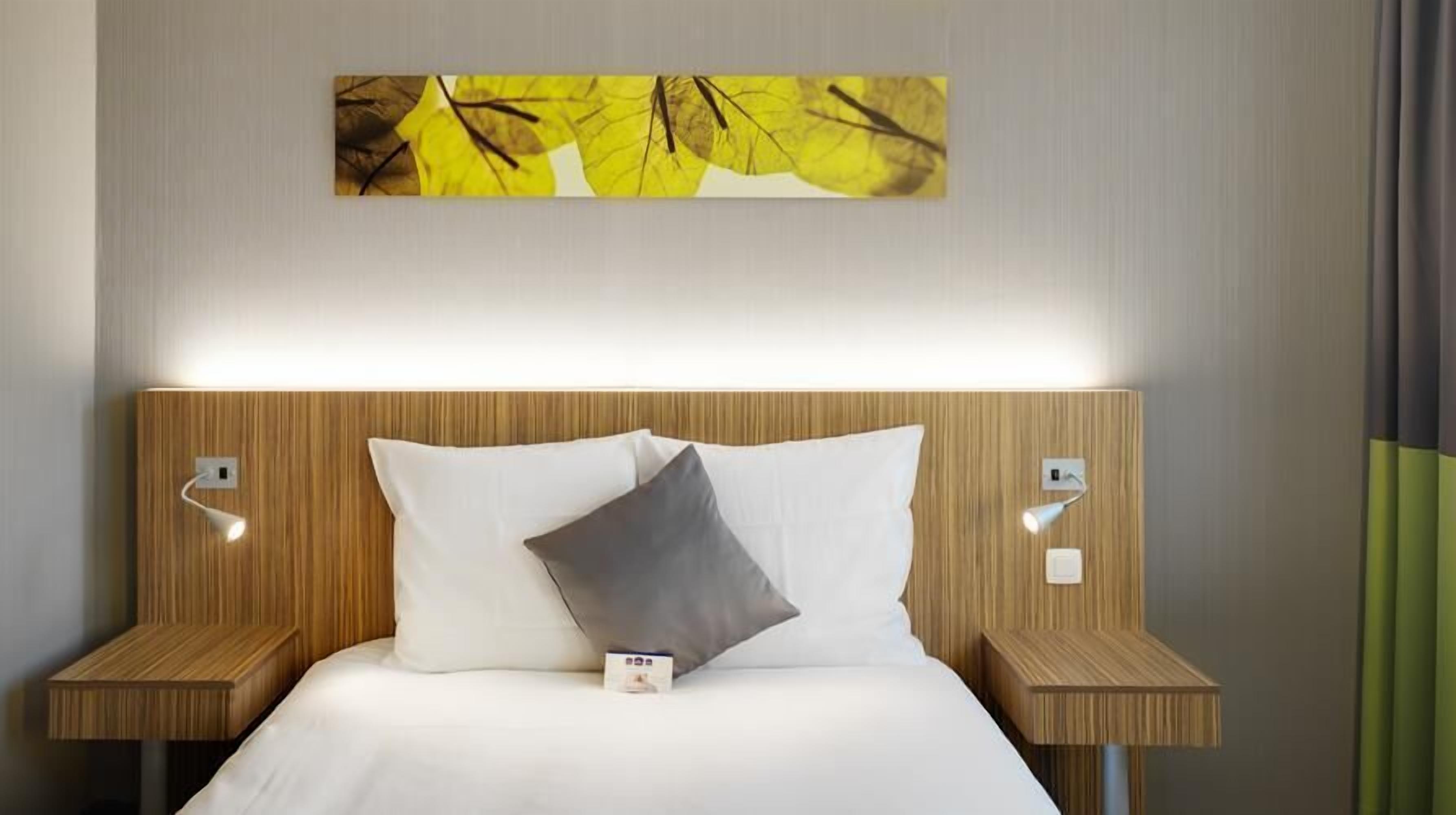 Una habitación de hotel con una cama, dos mesitas de noche, una lámpara de pared y un cuadro sobre la cama.