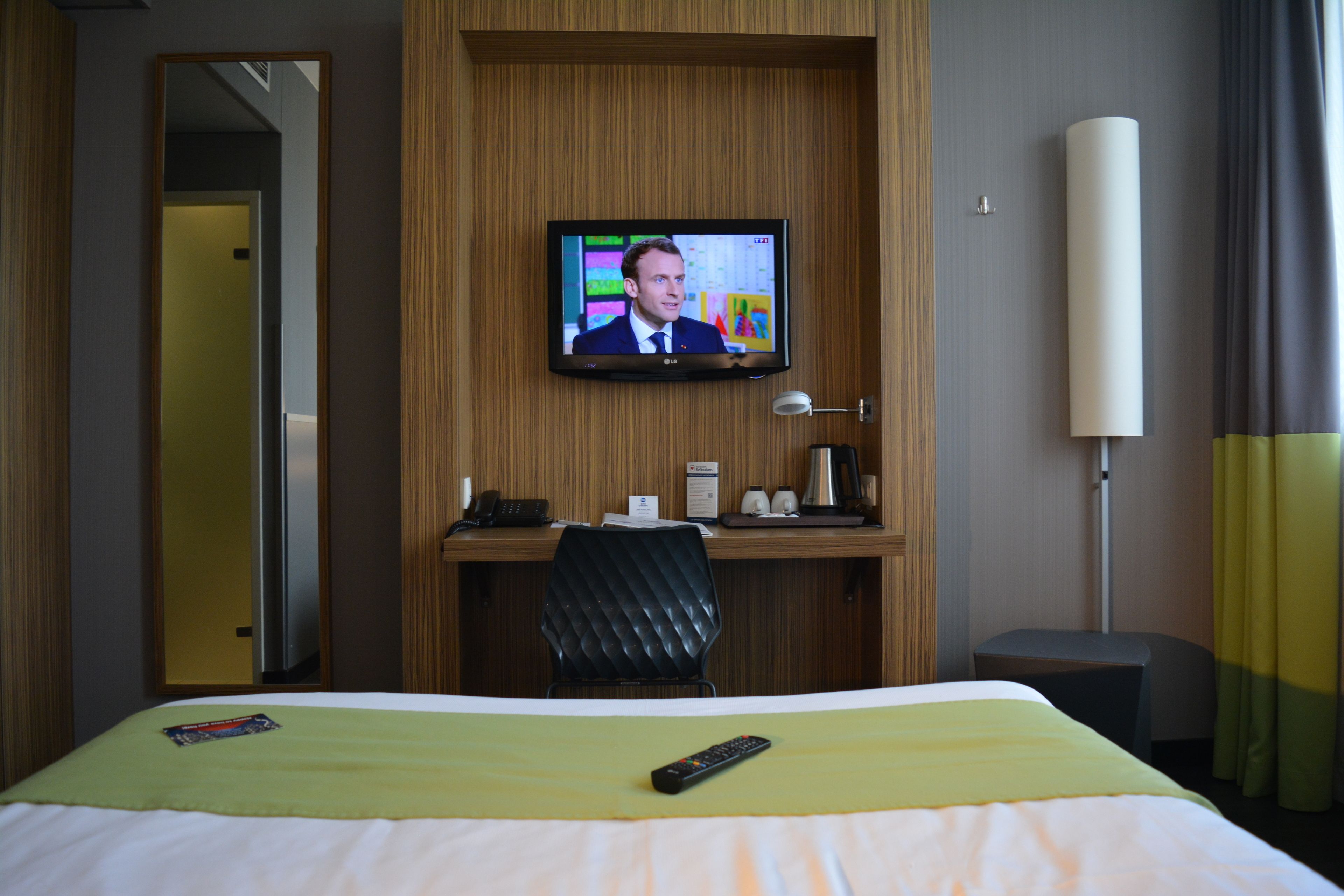 A hotel room with a bed, a desk with a computer, a TV mounted on the wall, and a bedside table with a lamp.