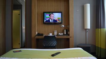 Quarto de hotel com cama, escrivaninha com computador, TV na parede e mesa de cabeceira com luminária.