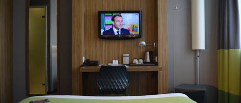 A hotel room with a bed, a desk with a computer, a TV mounted on the wall, and a bedside table with a lamp.