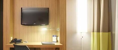Habitación de hotel con un escritorio de madera, una silla negra, un televisor montado en la pared, una mesita de noche y una ventana con cortinas.