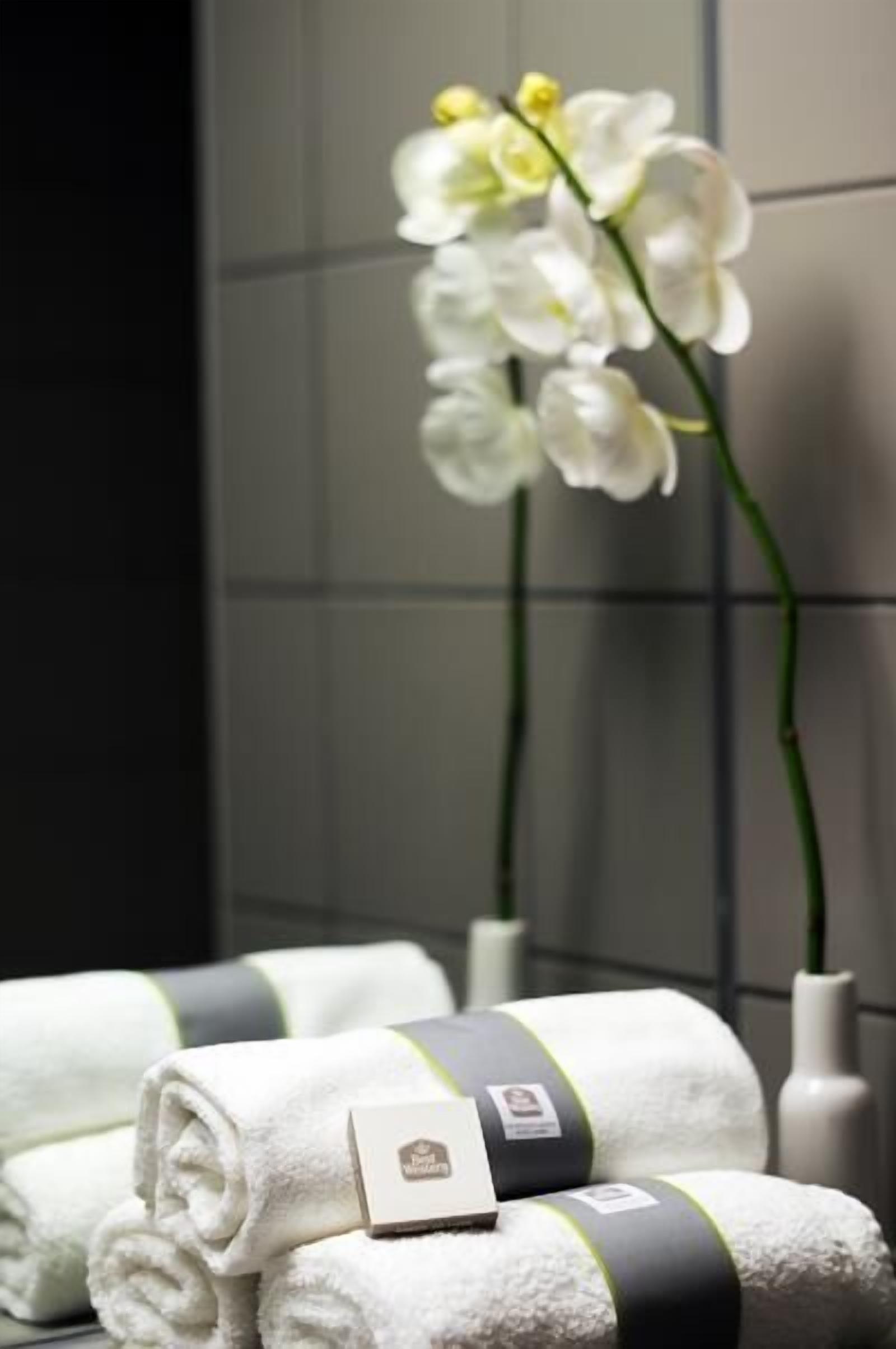 Un baño con toallas blancas, una orquídea blanca y un jarrón blanco.