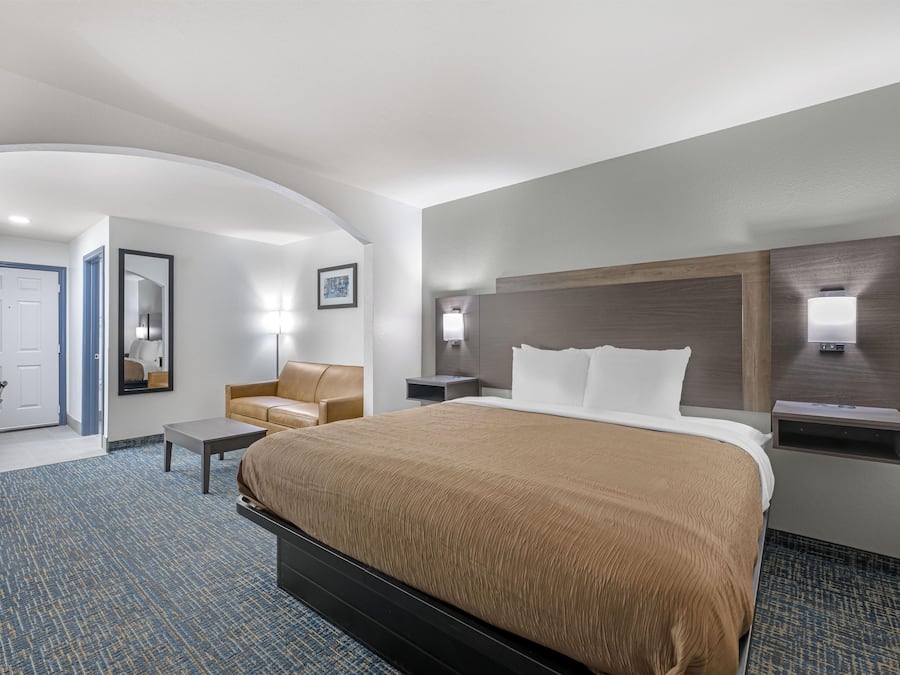 A hotel room with a large bed, a desk, a chair, a sofa, and a bathroom visible through an archway.