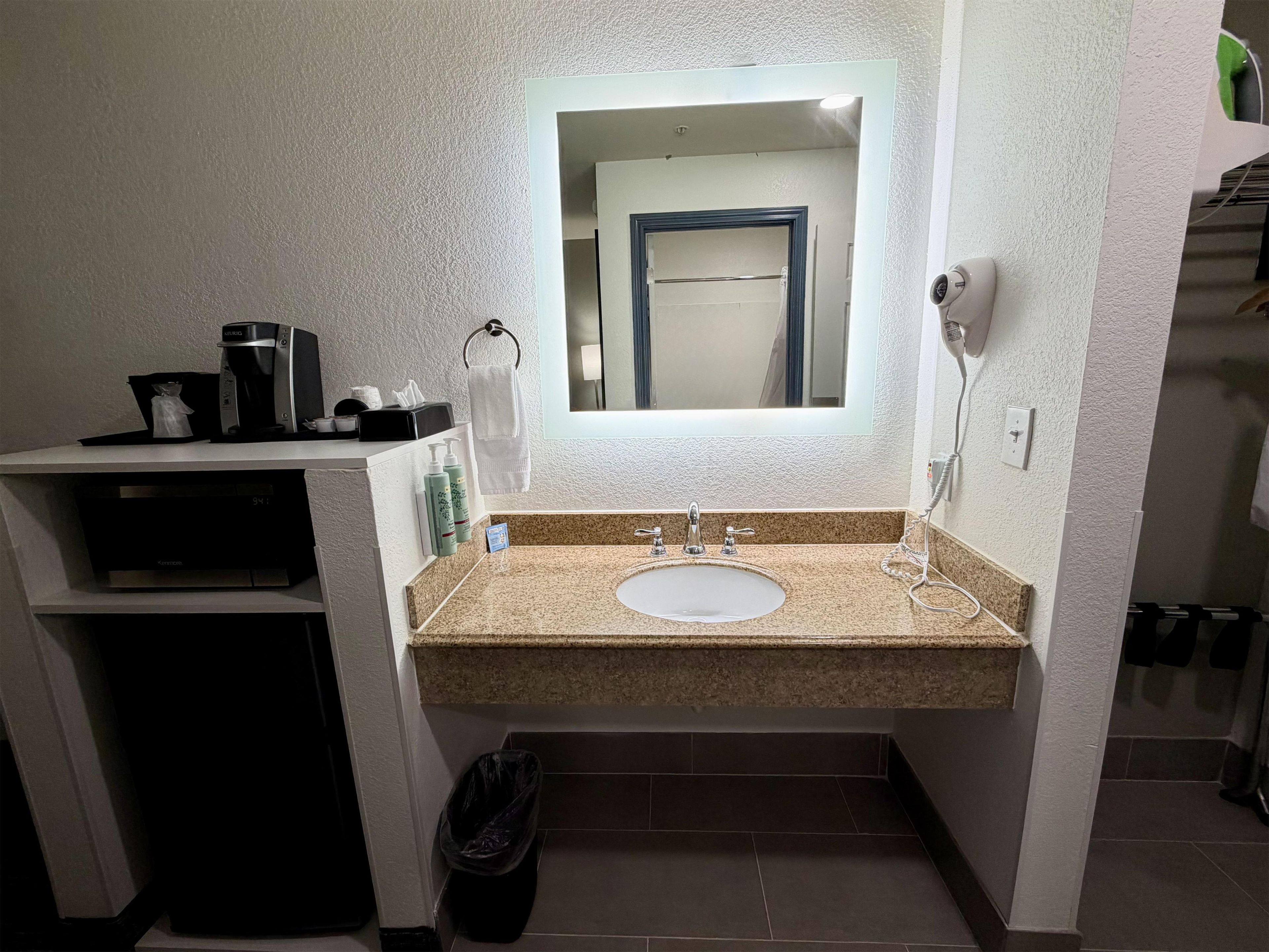Un baño con lavamanos, espejo, toallero y una cafetera en la encimera.