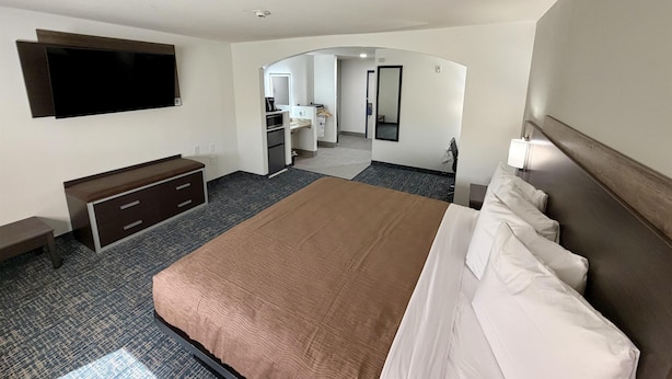 A hotel room with a large bed, a flat-screen TV mounted on the wall, a desk with a chair, and a bathroom visible through an archway.