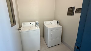 A laundry room with two white Whirlpool washers and dryers, a door with a mirror, and a wall-mounted sign indicating guest laundry services.