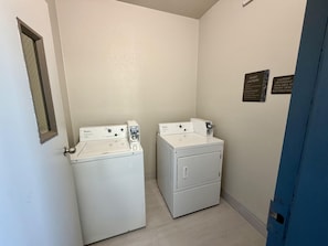 A laundry room with two white Whirlpool washers and dryers, a door with a mirror, and a wall-mounted sign indicating guest laundry services.