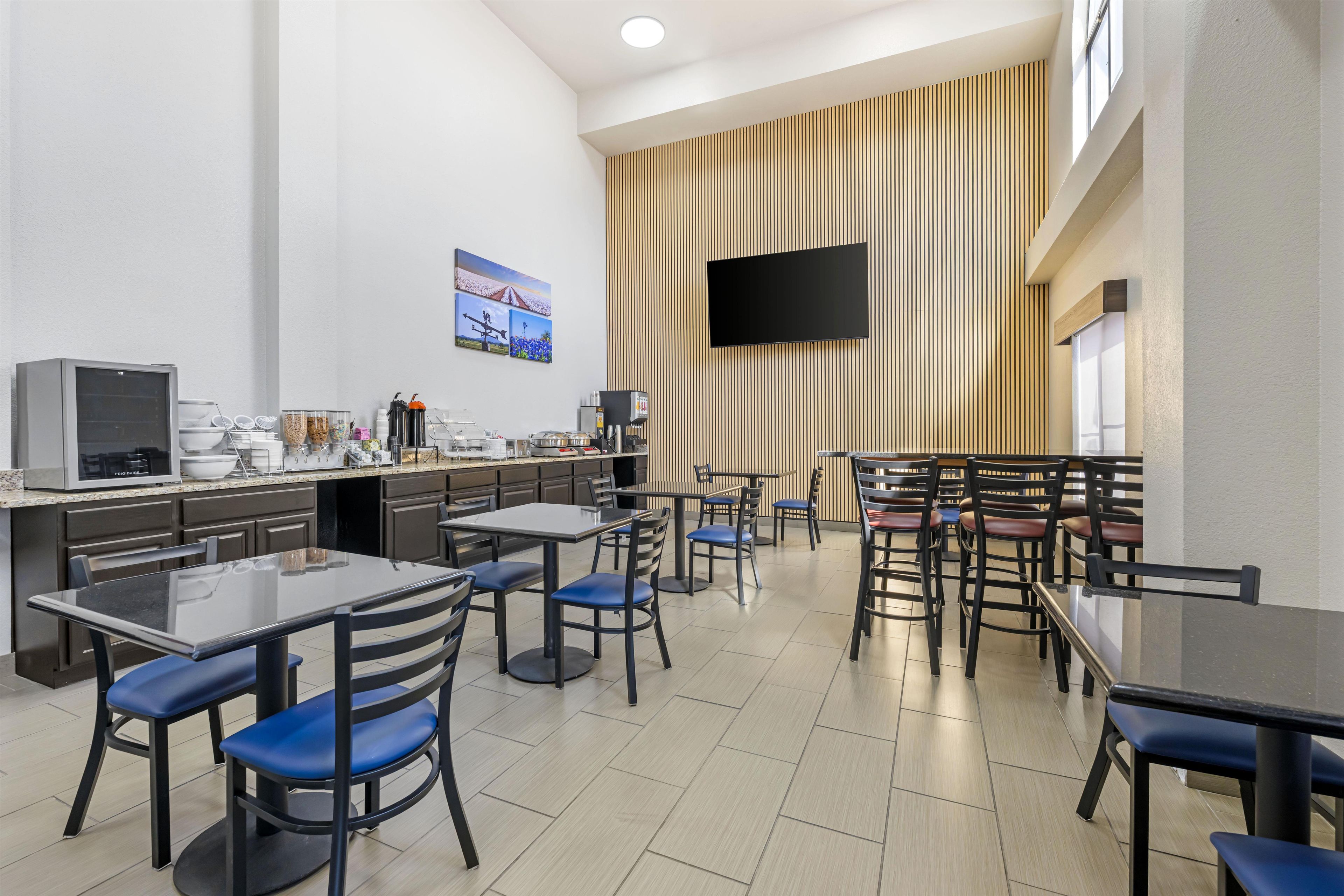 Un espace repas moderne avec des tables et des chaises, un coin cuisine équipé d’appareils électroménagers et un téléviseur à écran plat fixé au mur.