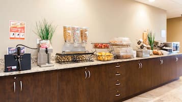 A hotel reception area with a coffee machine, juice dispensers, a fruit bowl, and a buffet setup.