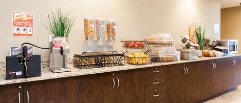 Zona de recepción de un hotel con una máquina de café, dispensadores de zumo, un frutero y un sistema de buffet.