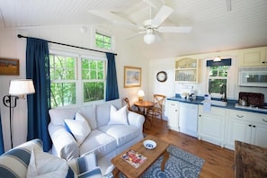 A cozy living room with a sofa, coffee table, and a dining area with a table and chairs. A kitchen with white cabinets and a microwave is visible.