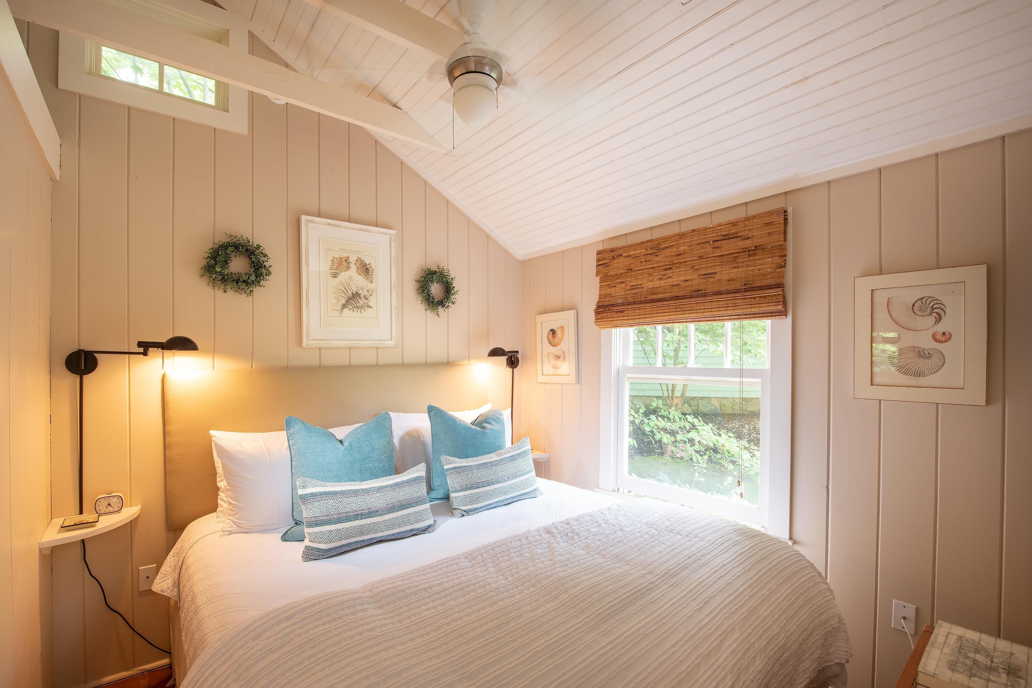 A bedroom with a bed, two wall-mounted lights, a window with a view, and two wall decorations.