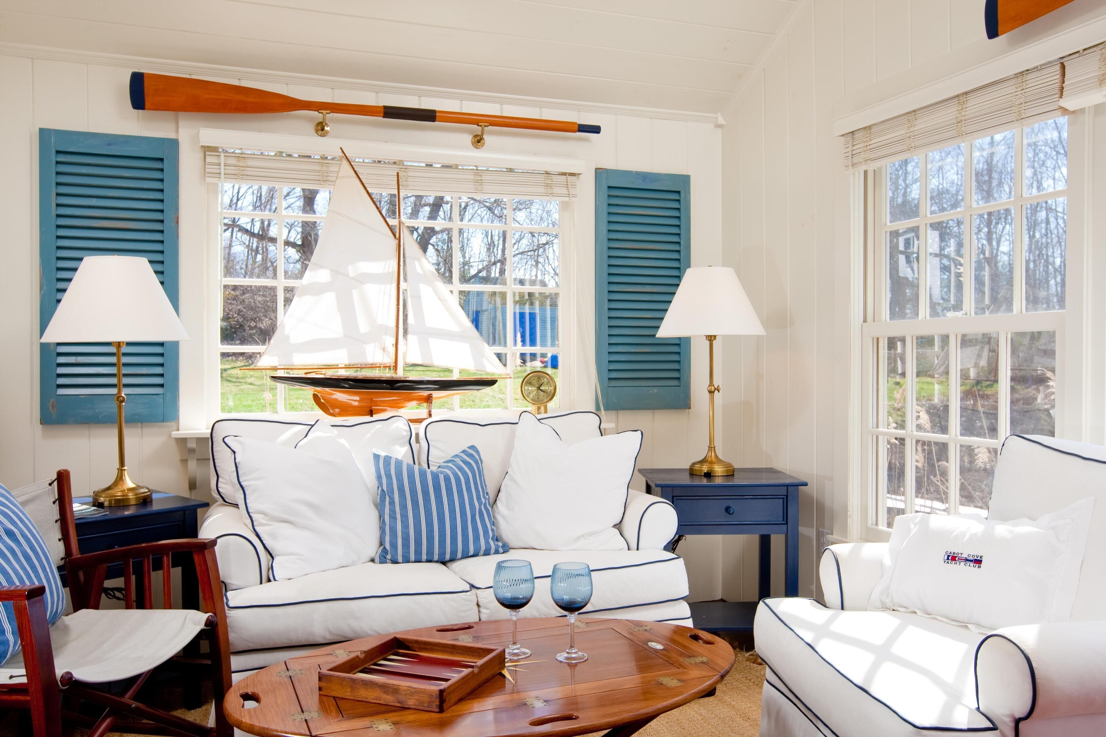 A cozy living room with a white sofa, blue and white pillows, a wooden coffee table with wine glasses, and a sailboat model.
