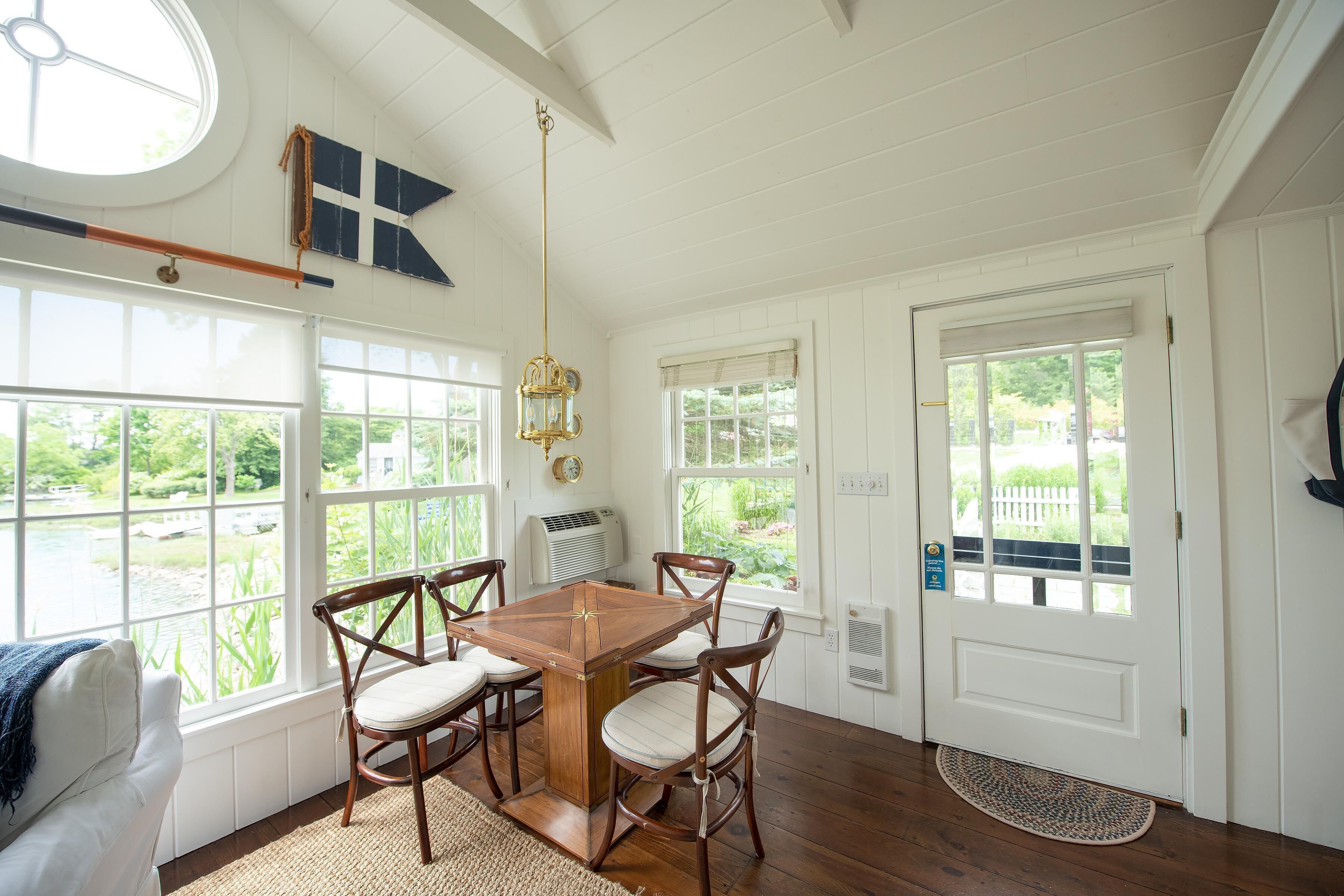 A dining area with a wooden table and chairs, large windows, and a view of greenery.