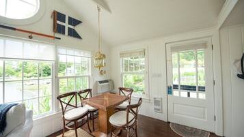 A dining area with a wooden table and chairs, large windows, and a view of greenery.