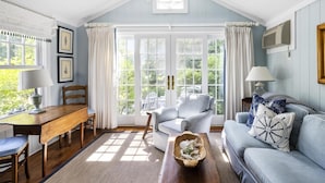 A cozy living room with a wooden dining table, a blue sofa, and a large window with curtains. - The Cottages at Cabot Cove (Kennebunkport)