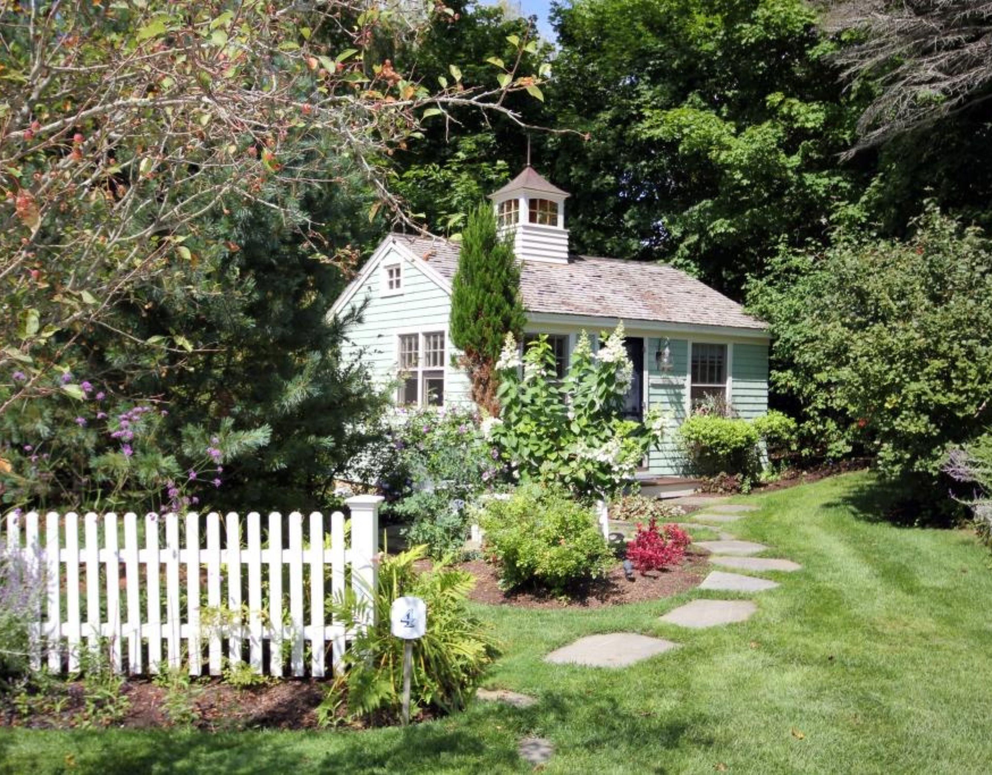 Une charmante maison avec une clôture en bois peinte en blanc, un jardin où poussent différentes espèces de plantes et un chemin de pierres menant à l’entrée.