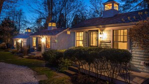 A two-story house with a well-lit front entrance, a visible chimney, and a small tower-like structure on the roof. - The Cottages at Cabot Cove (Kennebunkport)