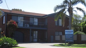A two-story brick building with a sign advertising a 2-bedroom air-conditioned unit, full kitchen & laundry facilities, solar heated pool & BBQ area, and undercover parking. - Harbour Lights Holiday Units (Merimbula)