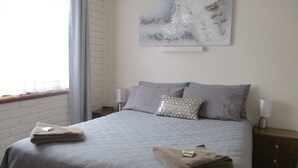A neatly made bed with a quilted cover, two pillows, and two folded towels on the bed. There is a wall-mounted artwork above the bed, a bedside table with a lamp, and a window with blinds. - Harbour Lights Holiday Units (Merimbula)