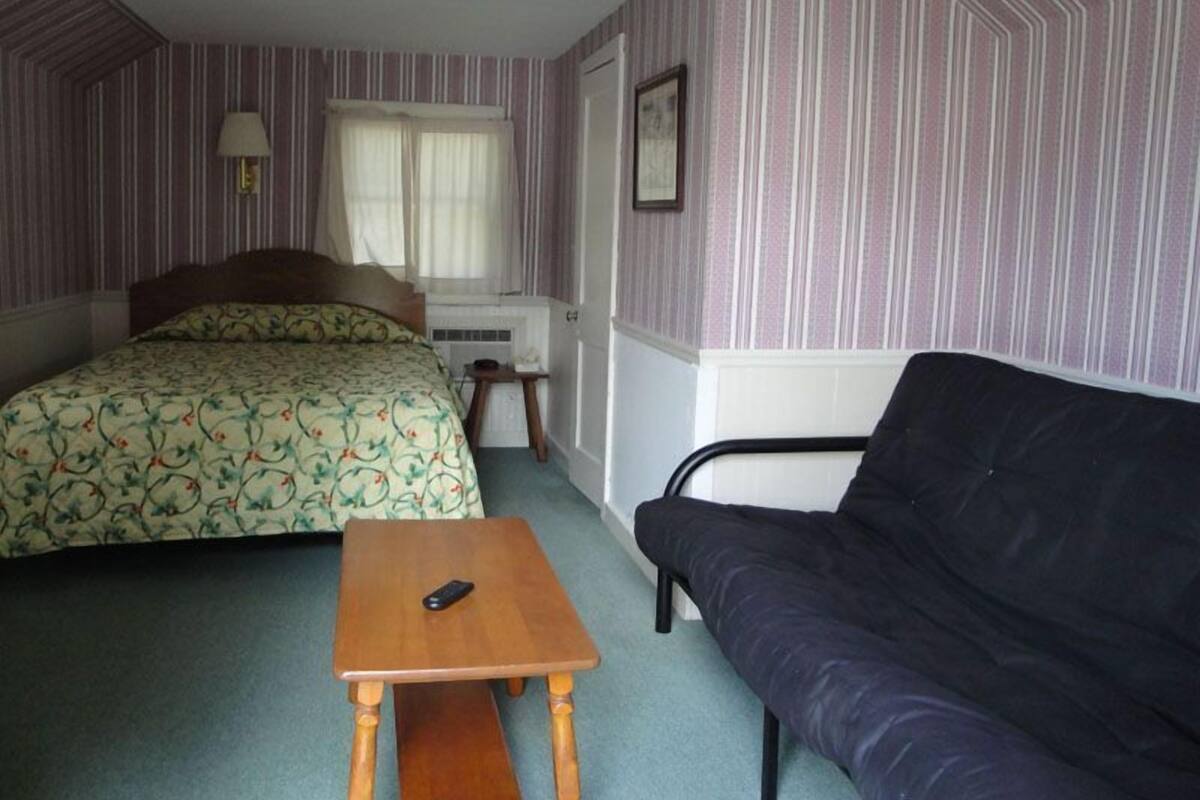 A bedroom with a bed, a small table, a sofa, a window with curtains, a lamp, and a radiator.