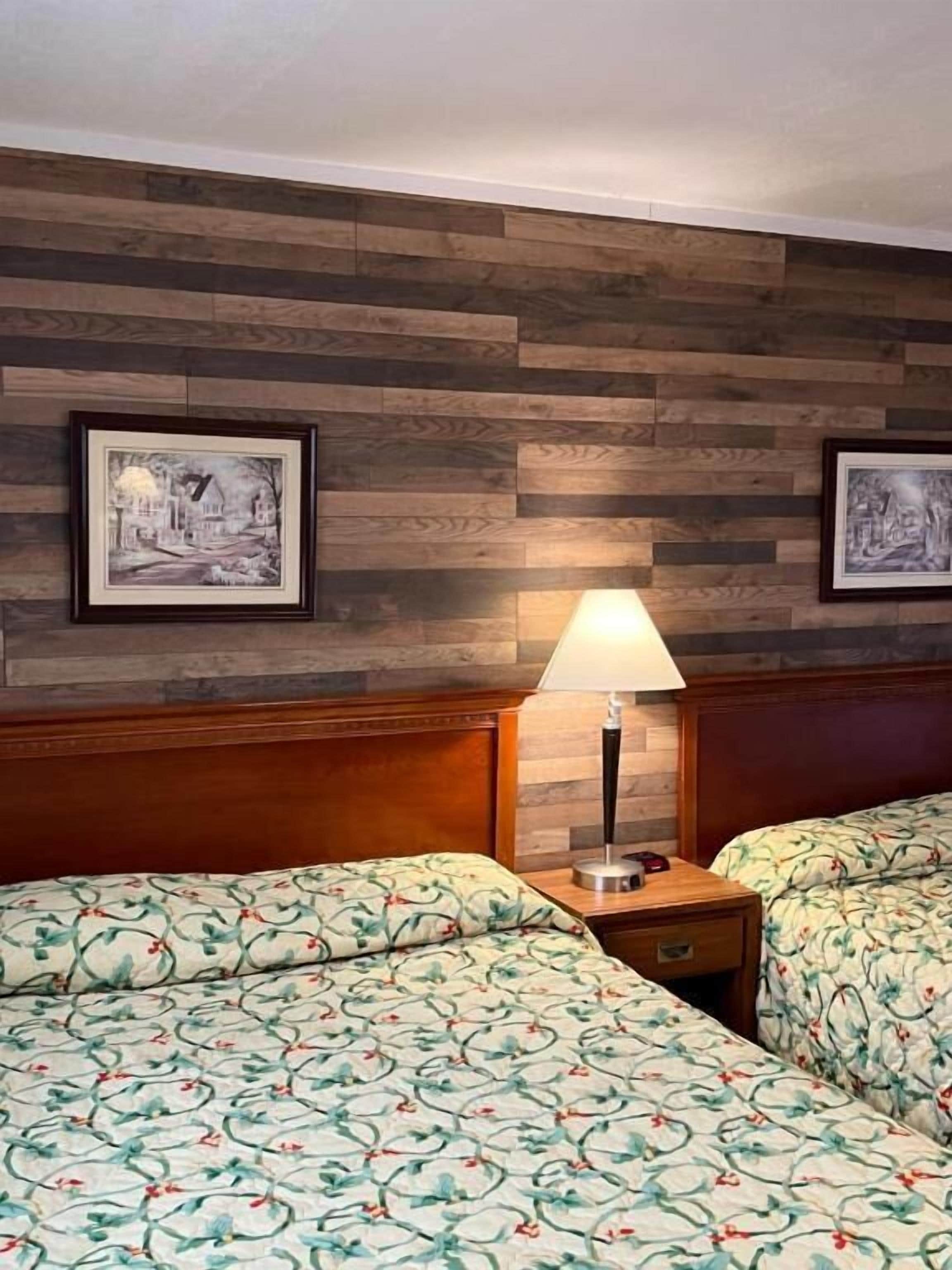 Habitación de hotel con dos camas, cabecera de madera, mesita de noche con lámpara y dos cuadros en la pared.