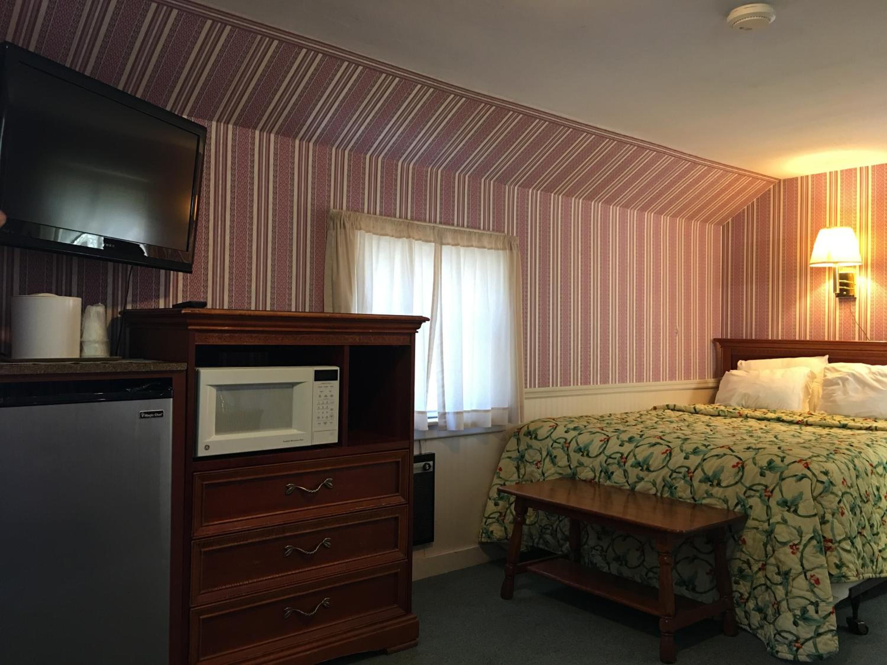 A hotel room with a bed, a wooden dresser, a microwave, a television, and a lamp.