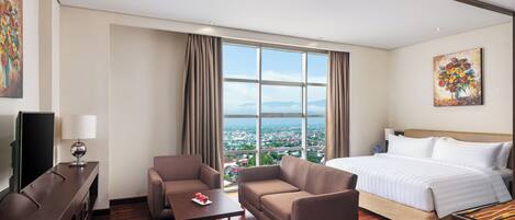 A hotel room with a large bed, a sofa, a coffee table, a TV, and a view of a cityscape through large windows.