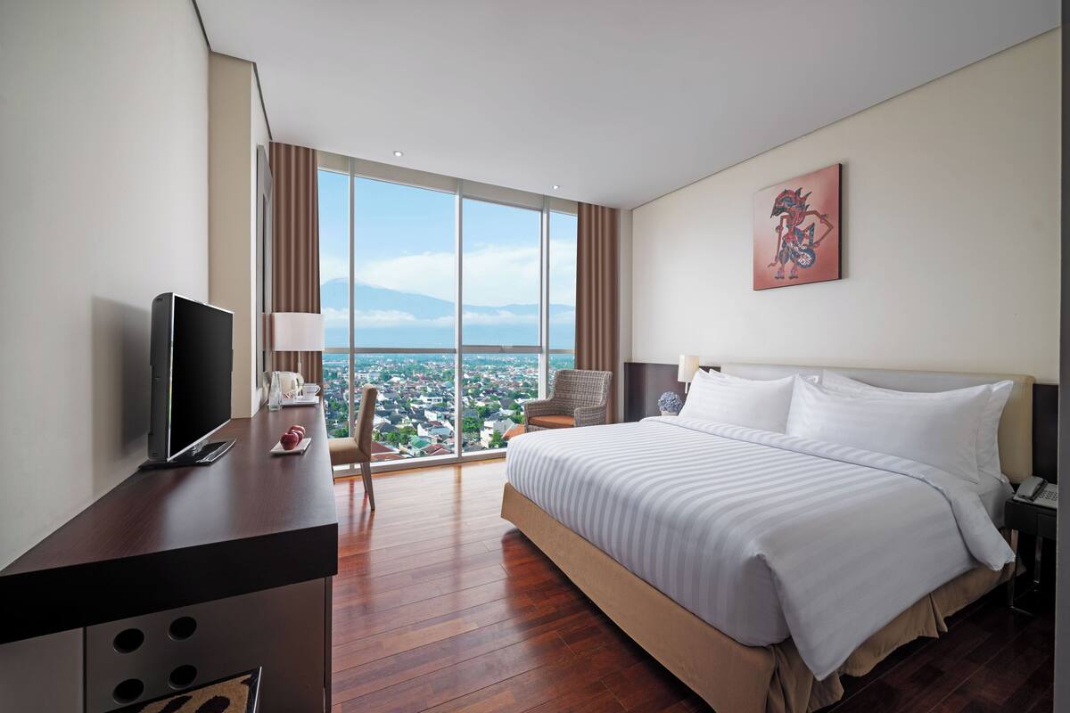Een hotelkamer met een groot bed, een bureau met een flatscreen-tv, een stoel en een groot raam met uitzicht op de stad.