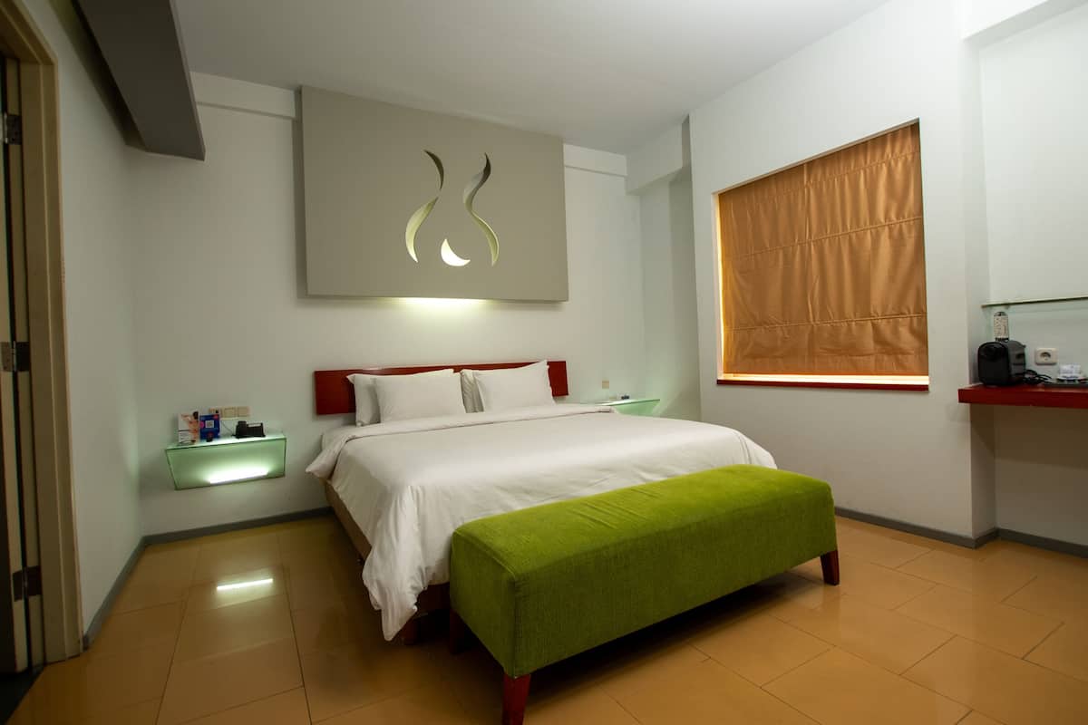 Une chambre d’hôtel avec un lit, un banc vert, une table de chevet, une œuvre d’art accrochée au mur et une fenêtre avec des stores.