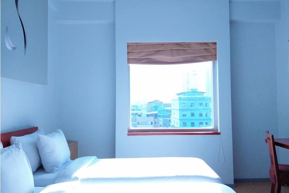Une chambre à coucher avec un lit, une fenêtre donnant sur des immeubles, une chaise et un mur au décor sobre.