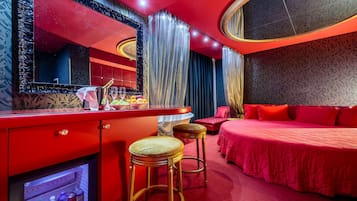 A hotel room with a red theme, including a bed, a vanity with a mirror, and a minibar.