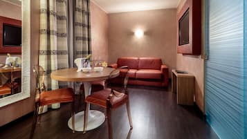 Una habitación de hotel con una mesa de comedor redonda, dos sillas, un sofá de cuero rojo, una pequeña mesita de noche y un televisor montado en la pared.