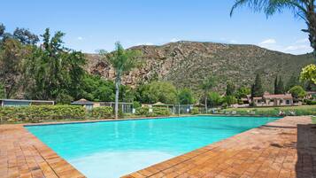 A swimming pool with a brick deck, surrounded by greenery and houses with a mountainous backdrop.