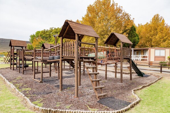 A wooden playground with climbing structures and a slide, surrounded by a gravel area and grass.