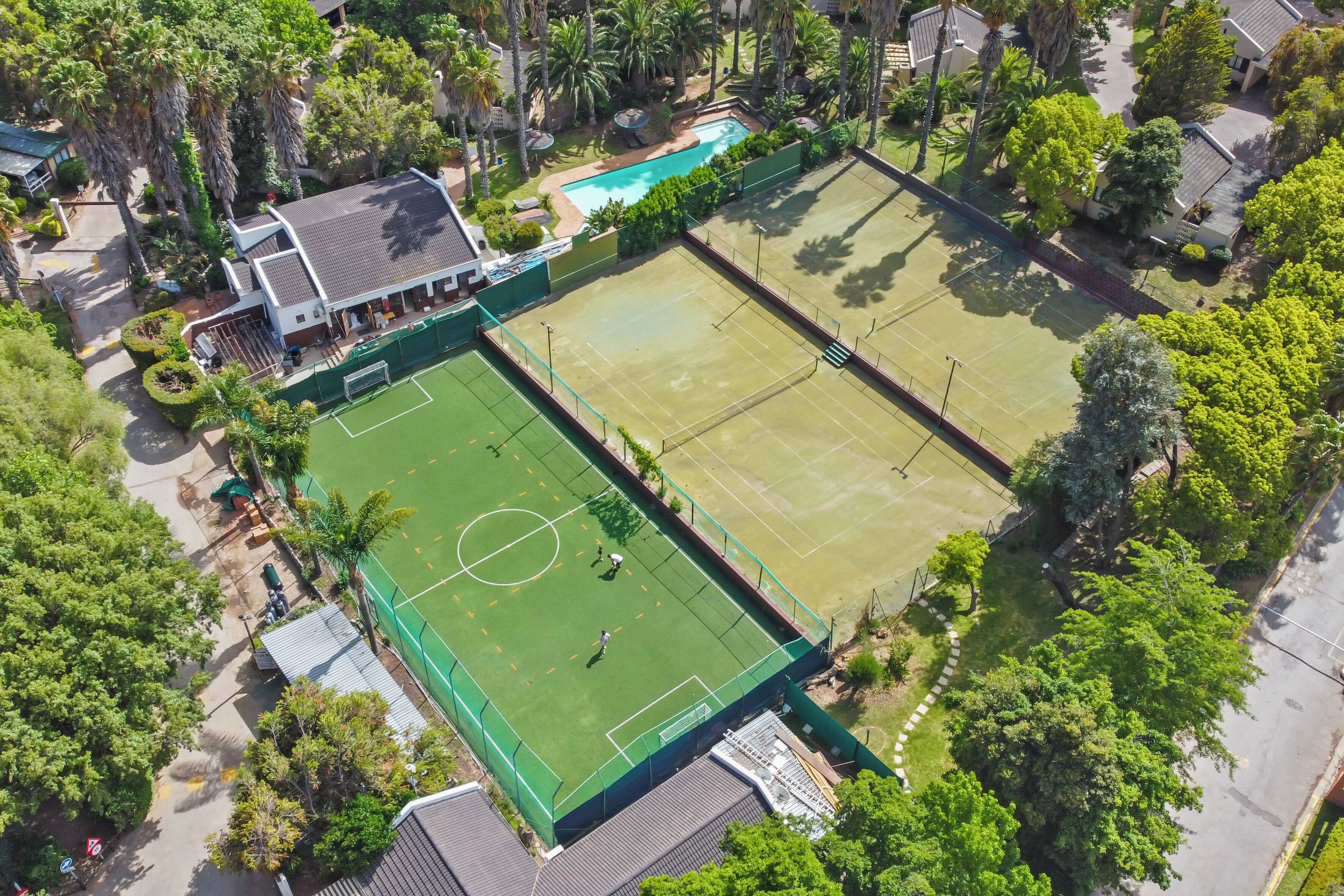 An aerial view of a residential area with two tennis courts, a swimming pool, and a building with a sloped roof.