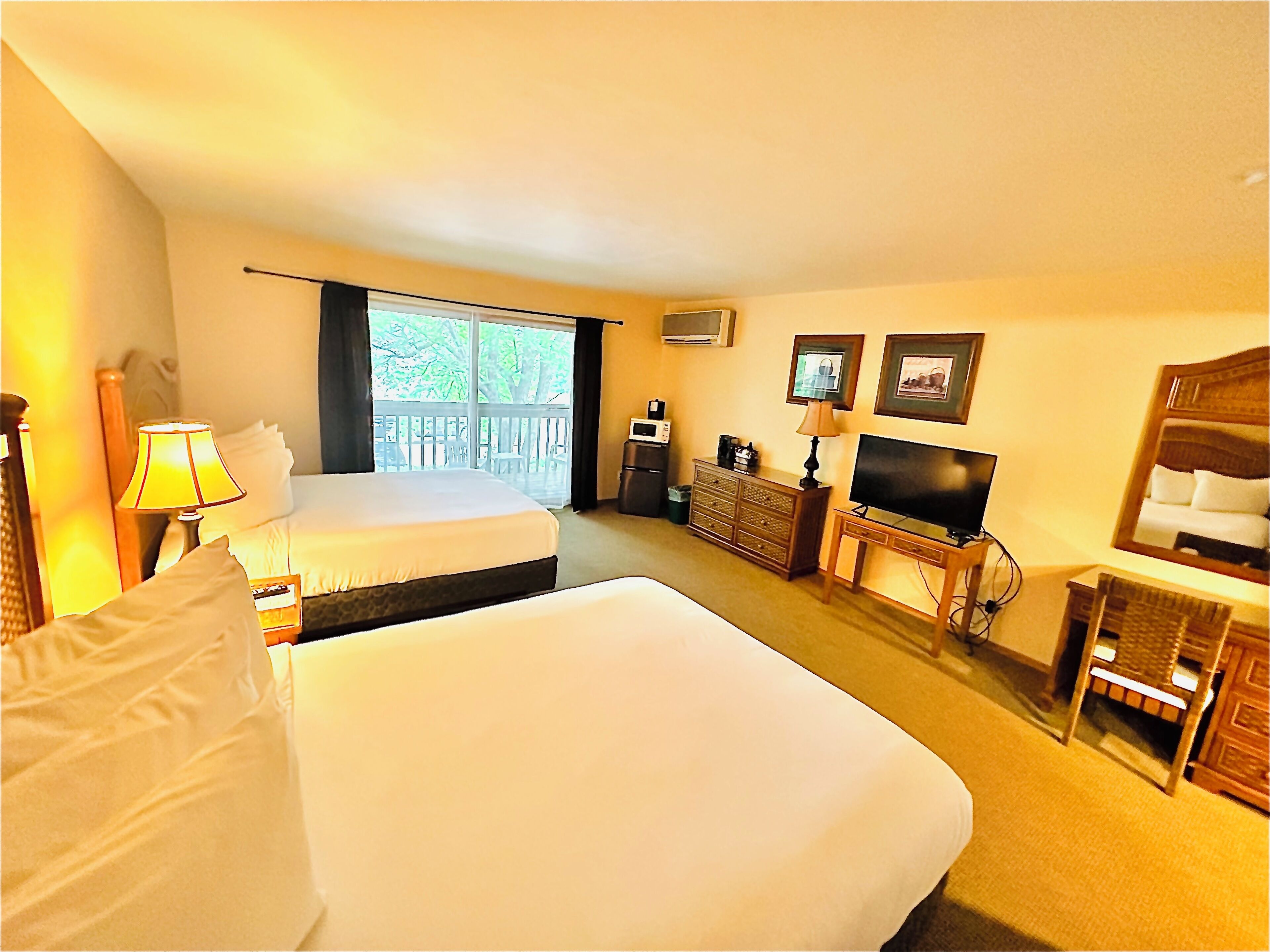 A hotel room with a large bed, a desk with a television, a chair, and a balcony with a view of trees.